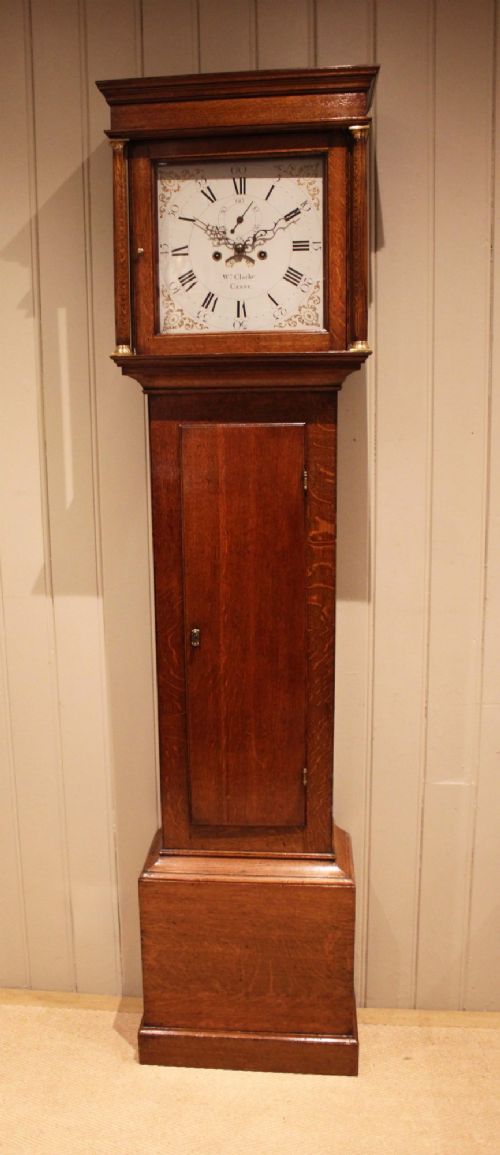A tall late Georgian oak longcase clock featuring a square painted dial, turned hood columns, and a polished wood trunk. View III