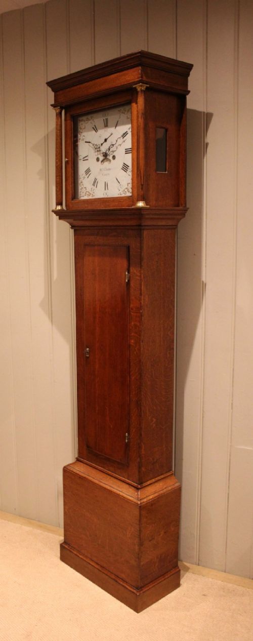 A tall late Georgian oak longcase clock featuring a square painted dial, turned hood columns, and a polished wood trunk. View IV