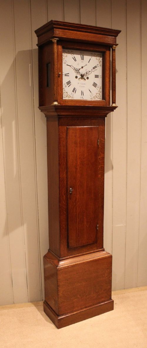 A tall late Georgian oak longcase clock featuring a square painted dial, turned hood columns, and a polished wood trunk. View V