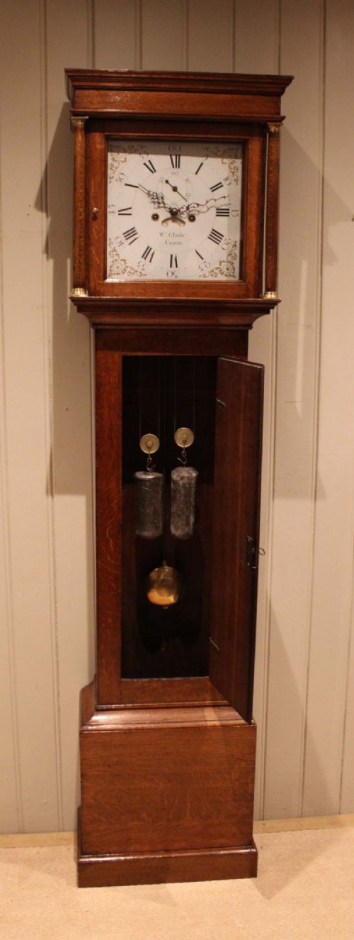 A tall late Georgian oak longcase clock featuring a square painted dial, turned hood columns, and a polished wood trunk. View XII