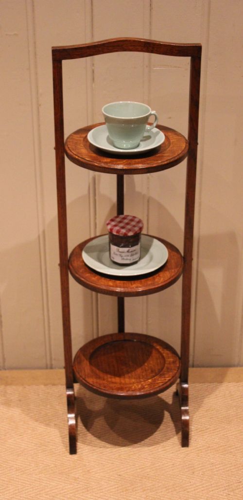 An Edwardian oak three-tier folding cake stand featuring circular recessed shelves and an arched carrying handle. View VI