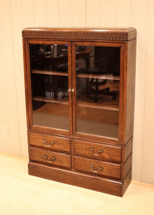fig 1. A medium brown oak glazed bookcase with two glass upper doors, carved egg-and-dart top moulding, and four drawers with brass handles.
