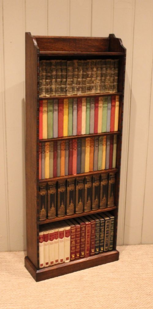 A tall, narrow dark oak open bookcase featuring five fixed shelves filled with vintage hardback books and a simple gallery top on a plinth base. View VII