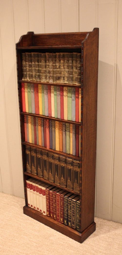 A tall, narrow dark oak open bookcase featuring five fixed shelves filled with vintage hardback books and a simple gallery top on a plinth base. View XI