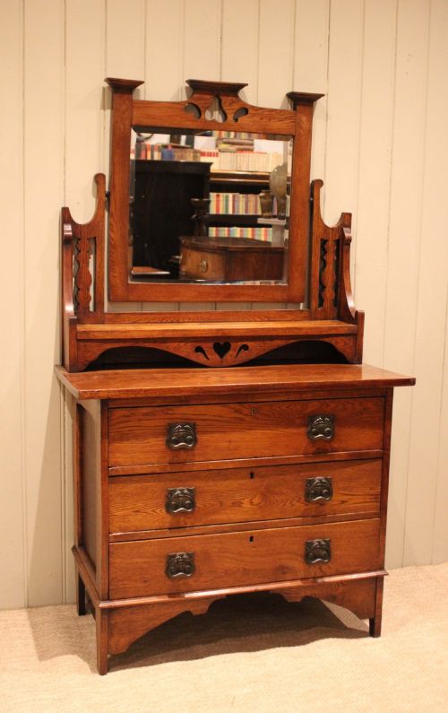 fig 1. A solid oak Arts and Crafts dressing chest featuring three wide drawers, metal drop handles, heart-shaped cutouts, and a tilting rectangular mirror on top.