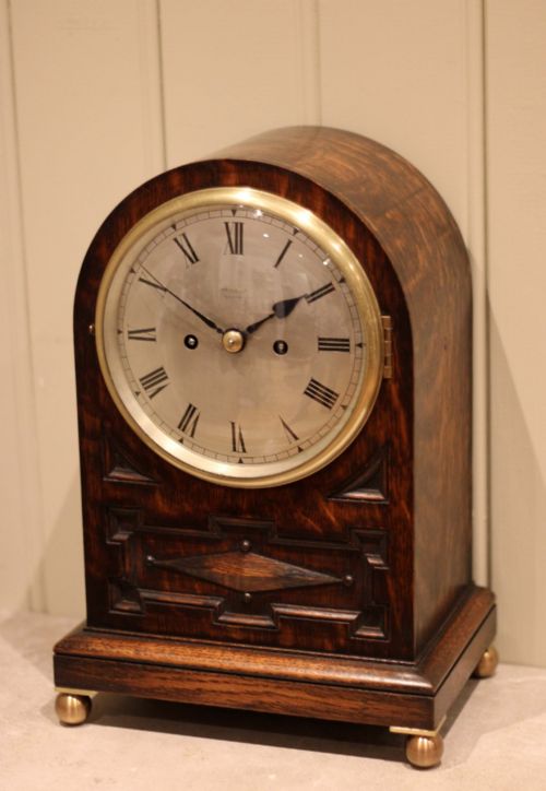 fig 2. A dark oak arched-top bracket clock featuring a circular silvered dial, brass bezel, geometric wooden moulding, and brass ball feet.