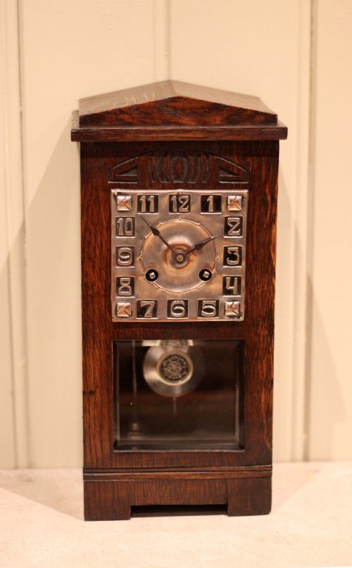 fig 1. An Arts and Crafts dark oak mantel clock with a pediment top, carved geometric detailing, a square silvered metal dial, and a lower glass aperture displaying the pendulum.