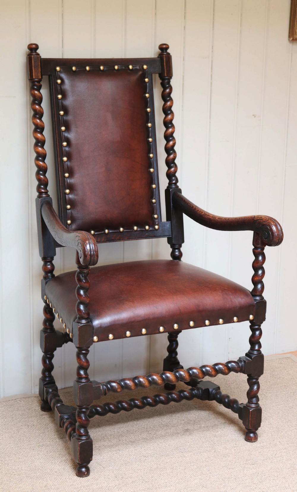 A high-backed dark oak armchair featuring barley twist columns, scroll armrests, and reddish-brown leather upholstery with brass studding.