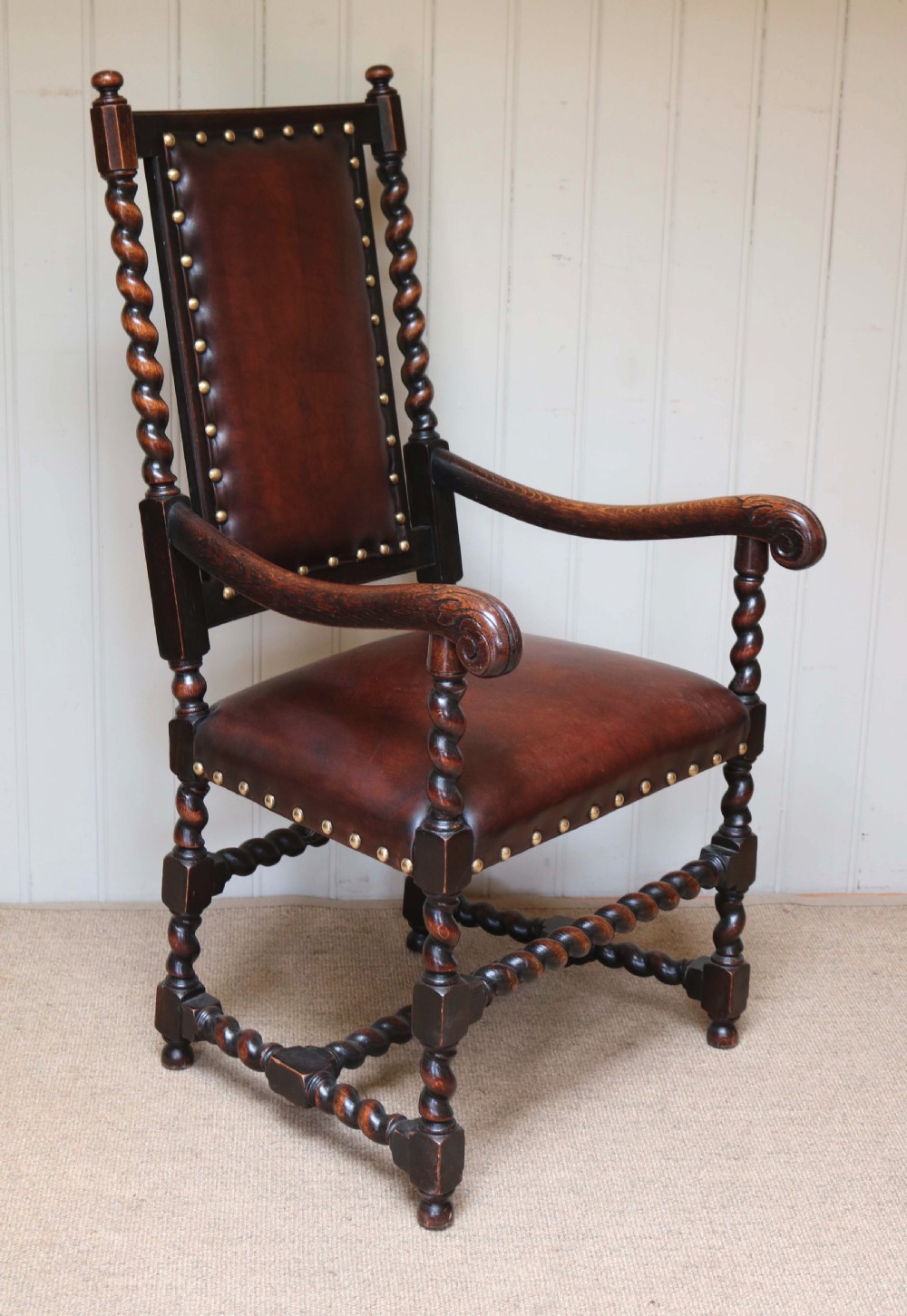 A high-backed dark oak armchair featuring barley twist columns, scroll armrests, and reddish-brown leather upholstery with brass studding. View V