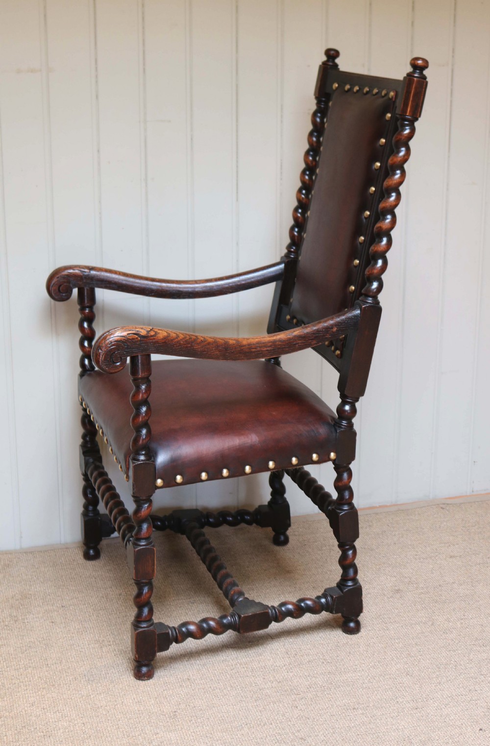 A high-backed dark oak armchair featuring barley twist columns, scroll armrests, and reddish-brown leather upholstery with brass studding. View VI