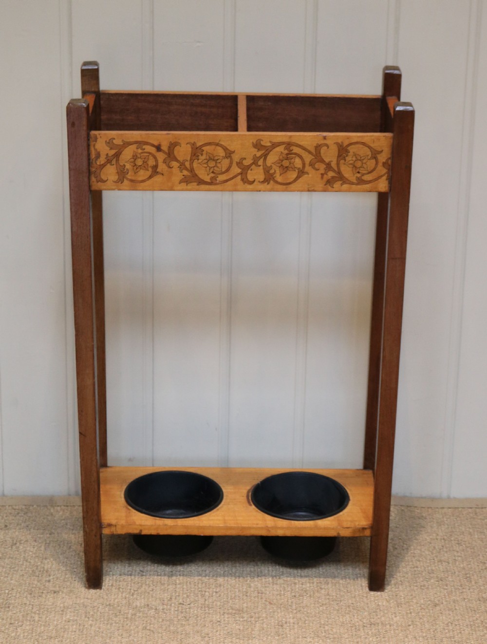 A dark wooden umbrella stand featuring a light inlaid maple top panel decorated with floral motifs, divided upper sections, and two circular black drip trays at the base.