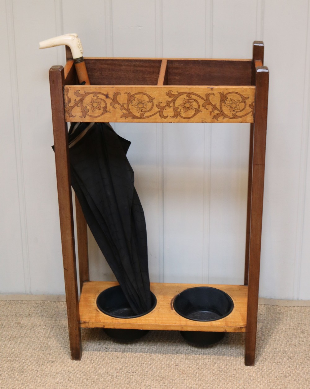 A dark wooden umbrella stand featuring a light inlaid maple top panel decorated with floral motifs, divided upper sections, and two circular black drip trays at the base. View III