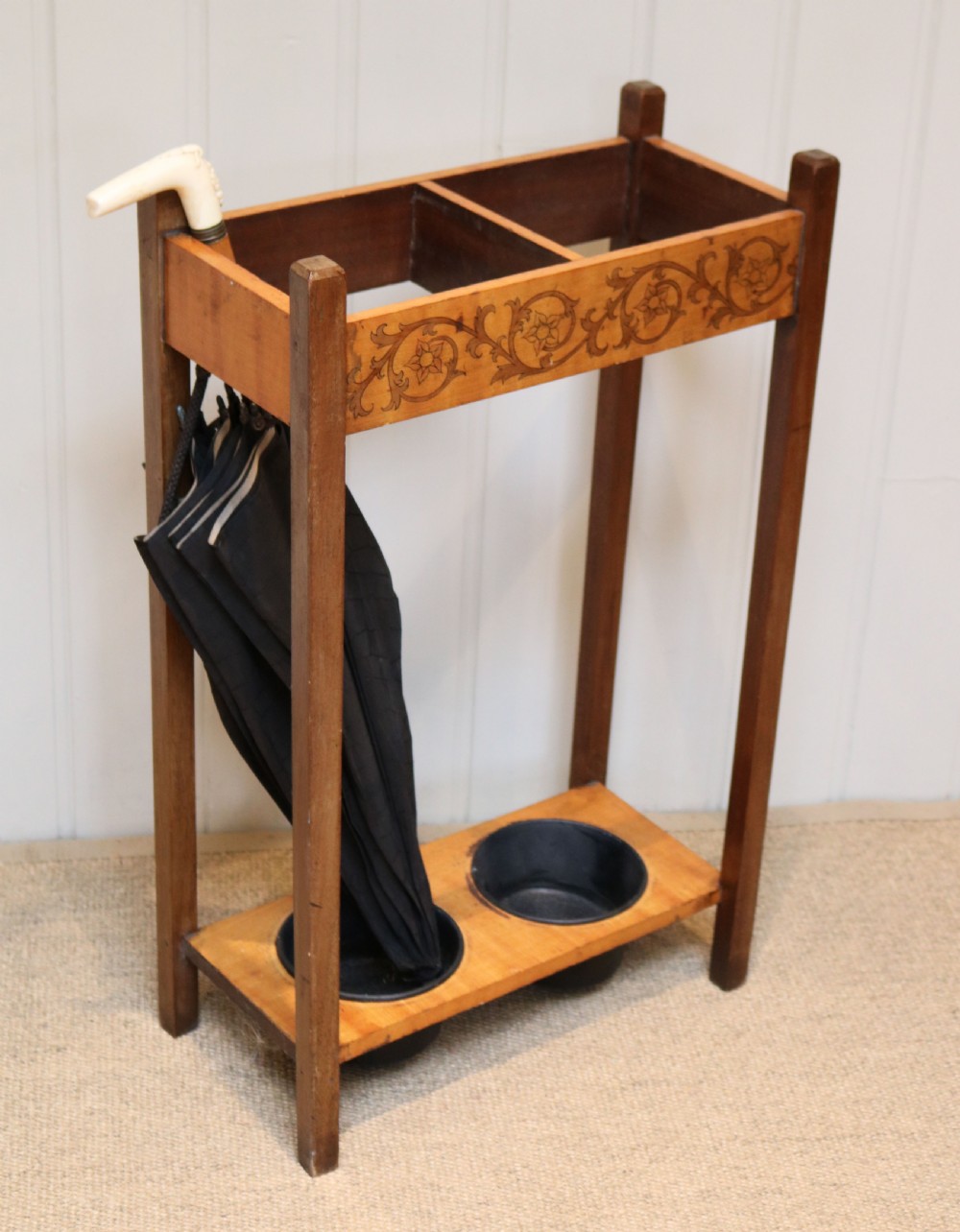 A dark wooden umbrella stand featuring a light inlaid maple top panel decorated with floral motifs, divided upper sections, and two circular black drip trays at the base. View V