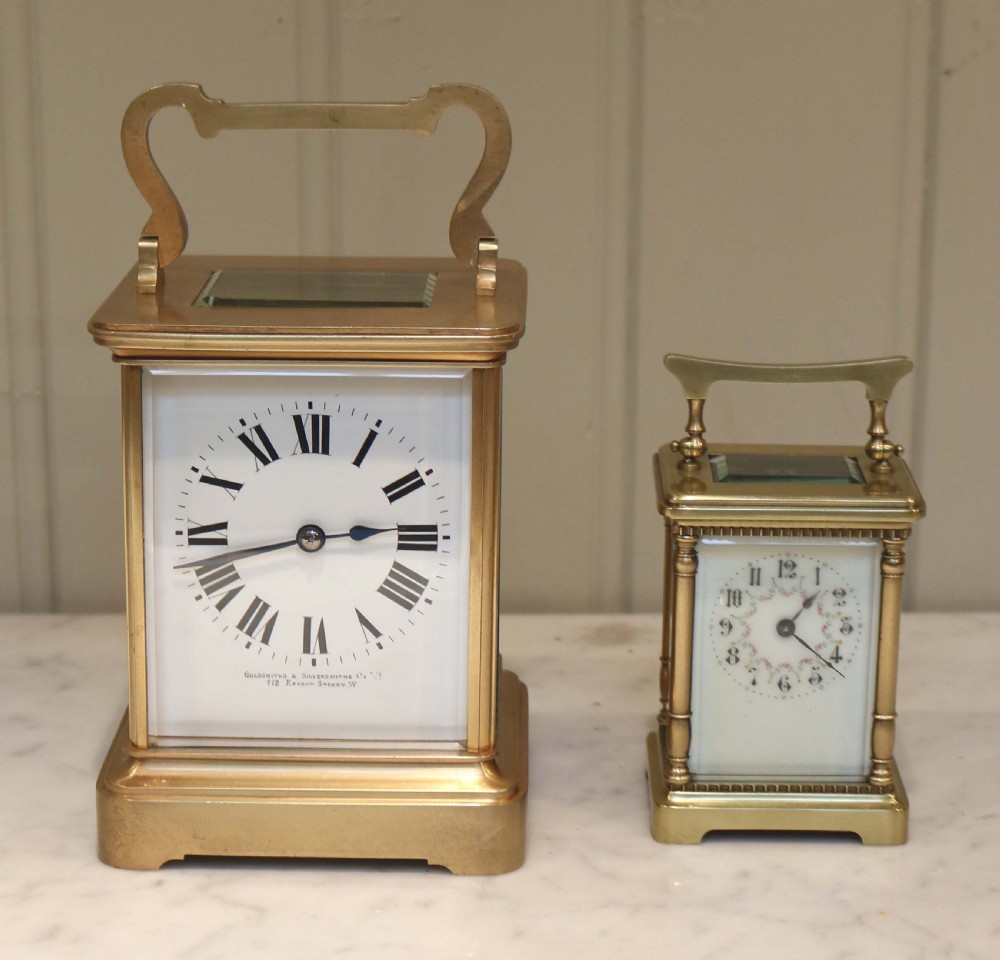 A large rectangular brass carriage clock featuring bevelled glass side panels, a white enamel dial with black Roman numerals, and a hinged top handle. View VI