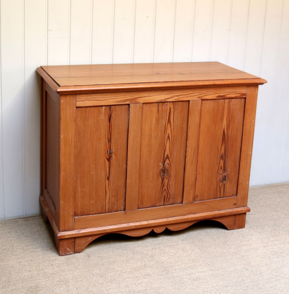 A rectangular warm pine storage box featuring three panelled front sections, a flat hinged top, and a shaped plinth base.