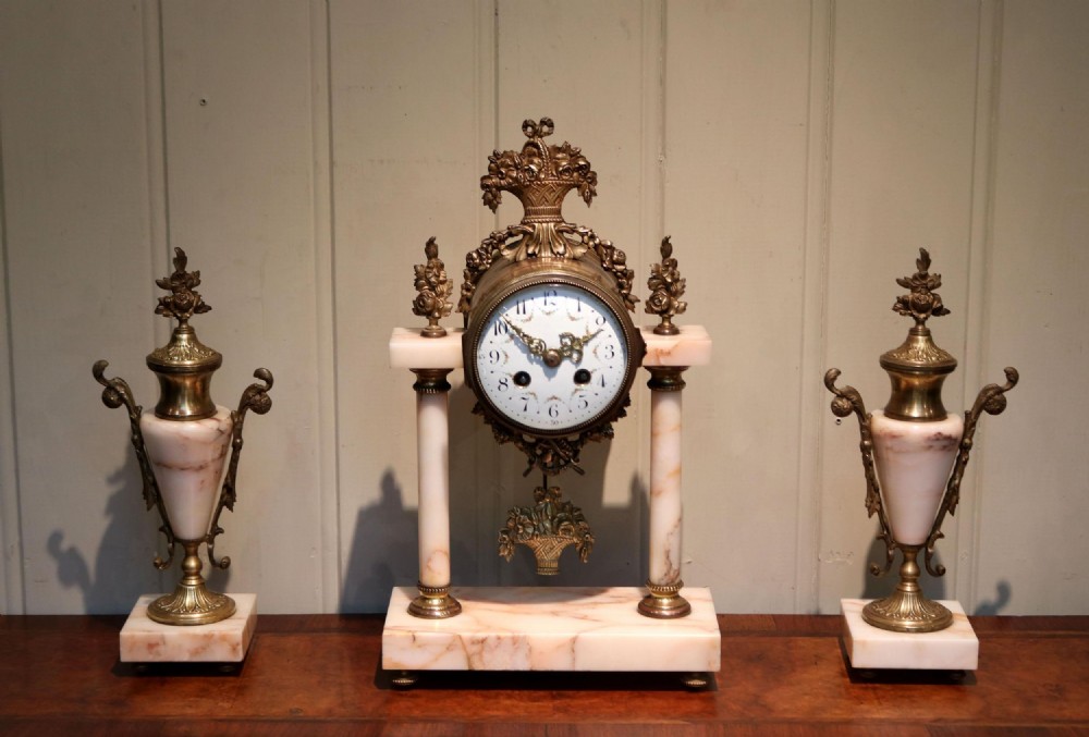 A three-piece French clock garniture set constructed from pale pink marble and gilt brass, featuring a central portico clock with a white enamel dial flanked by two matching decorative urns.