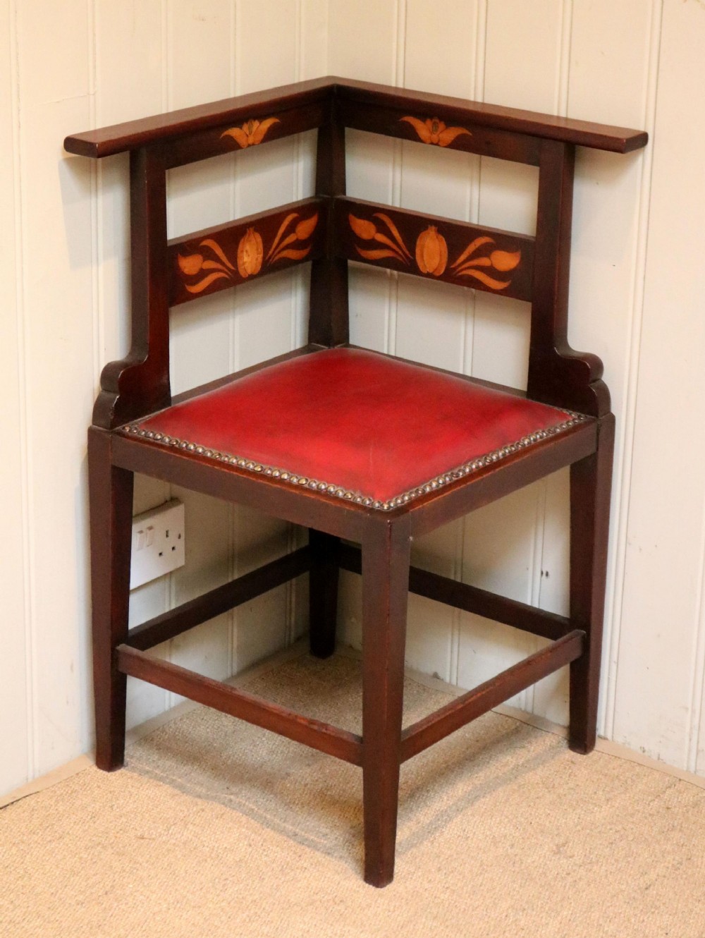 An Art Nouveau mahogany corner chair with floral marquetry inlay on the backrest and a red leather seat with brass stud detailing.