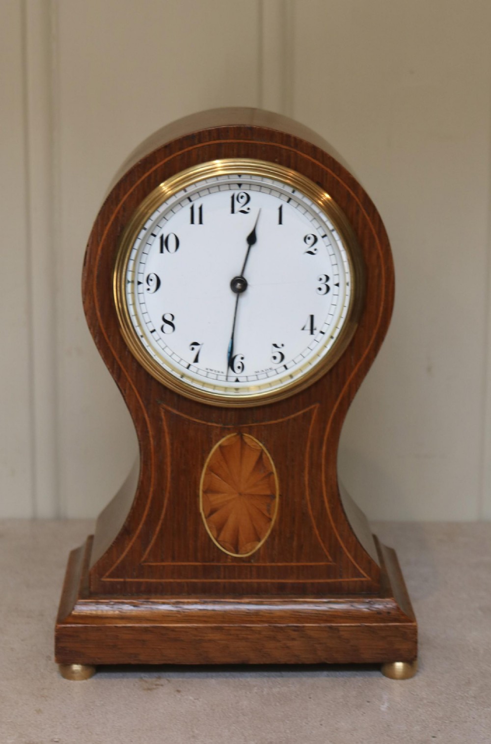 An Edwardian oak balloon mantel clock featuring an oval marquetry patera inlay, white enamel dial with Arabic numerals, brass bezel, and small brass feet.