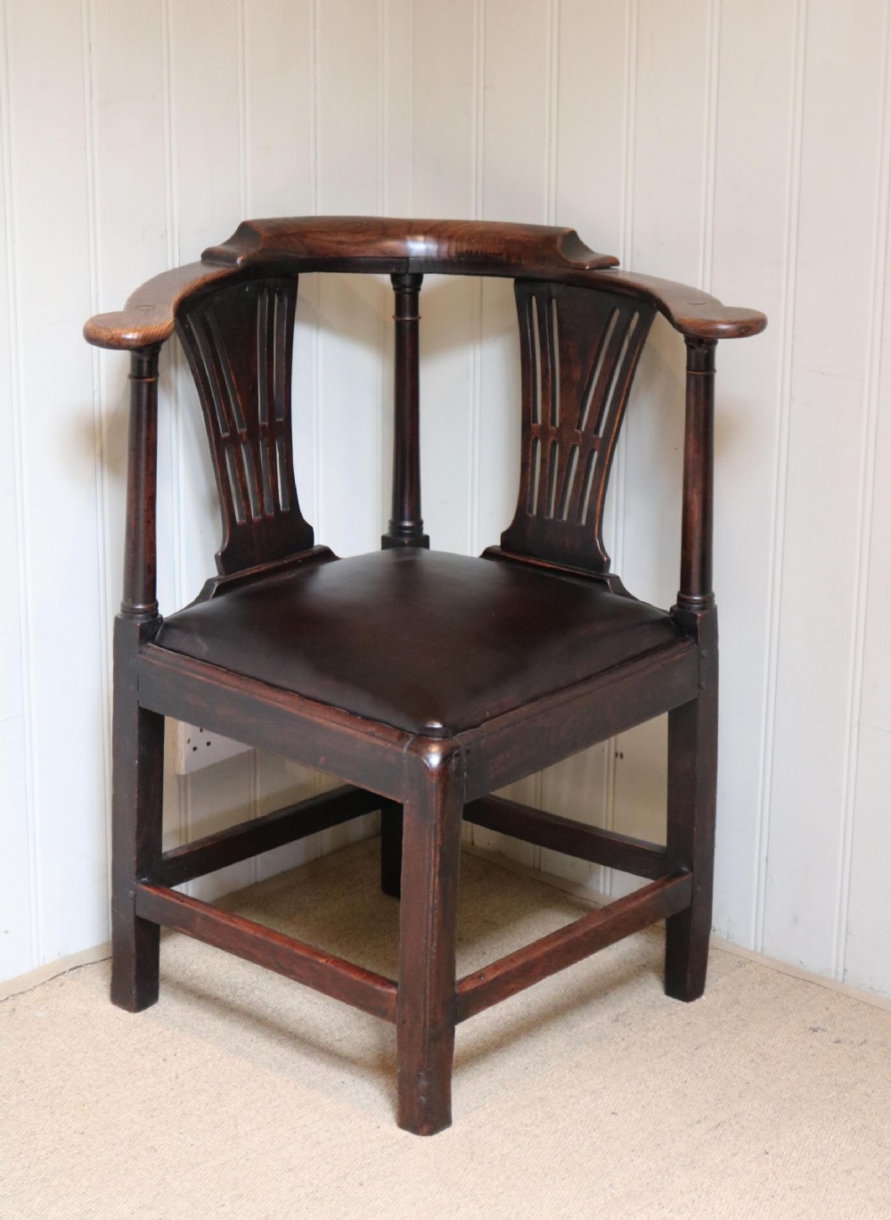 An antique dark elm corner chair featuring a curved armrest rail, twin pierced back splats, turned supports, and a dark upholstered seat.