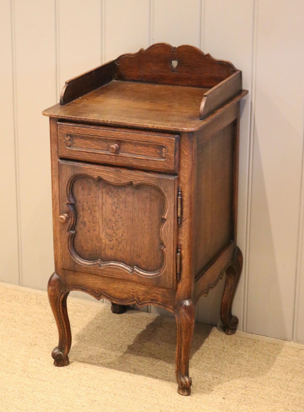 A warm brown solid oak bedside cabinet featuring a heart cut-out gallery, a single drawer, a carved panel door, and cabriole legs.