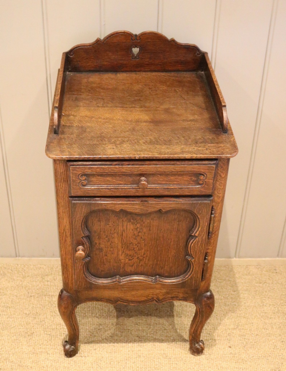 A warm brown solid oak bedside cabinet featuring a heart cut-out gallery, a single drawer, a carved panel door, and cabriole legs. View II