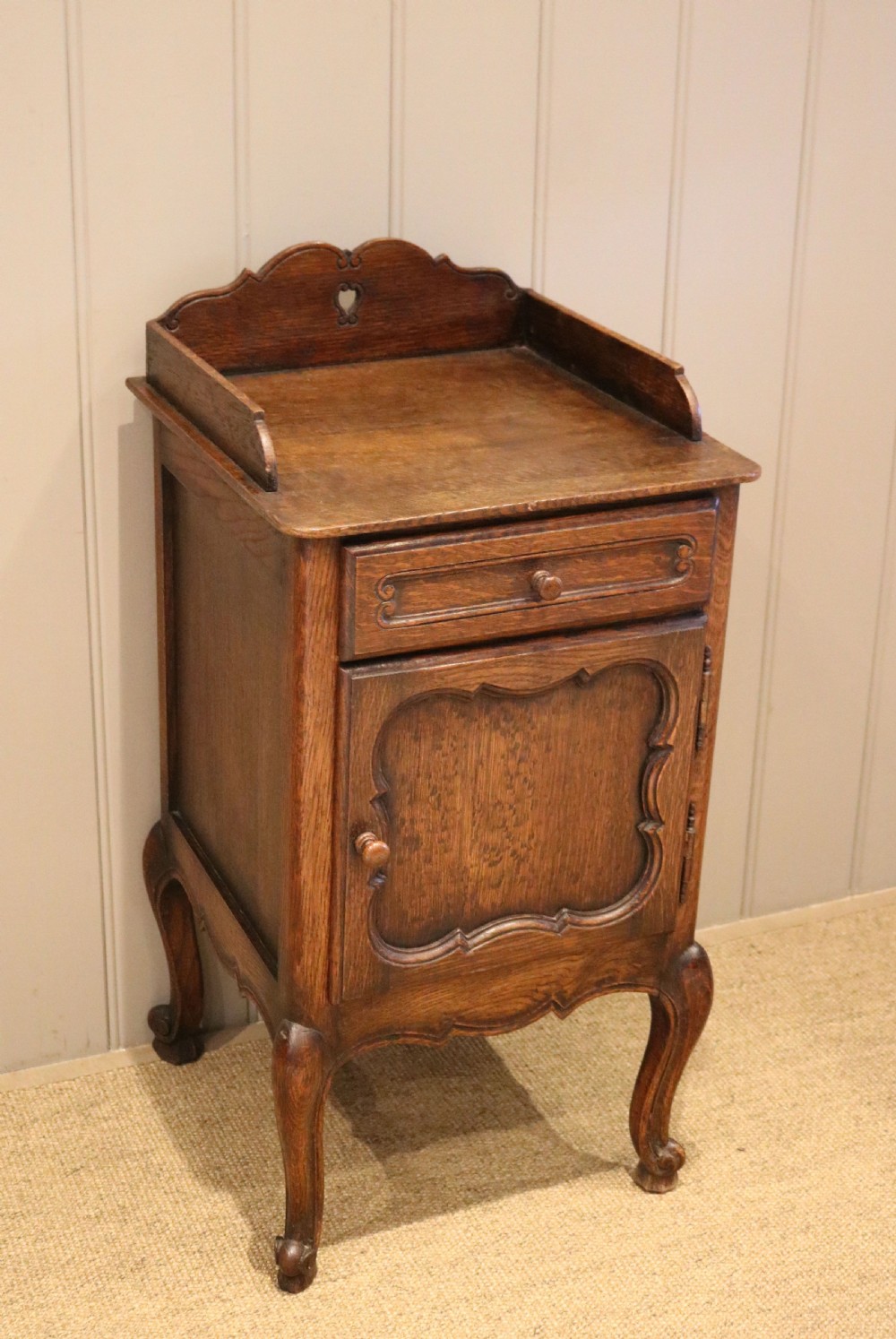 A warm brown solid oak bedside cabinet featuring a heart cut-out gallery, a single drawer, a carved panel door, and cabriole legs. View III