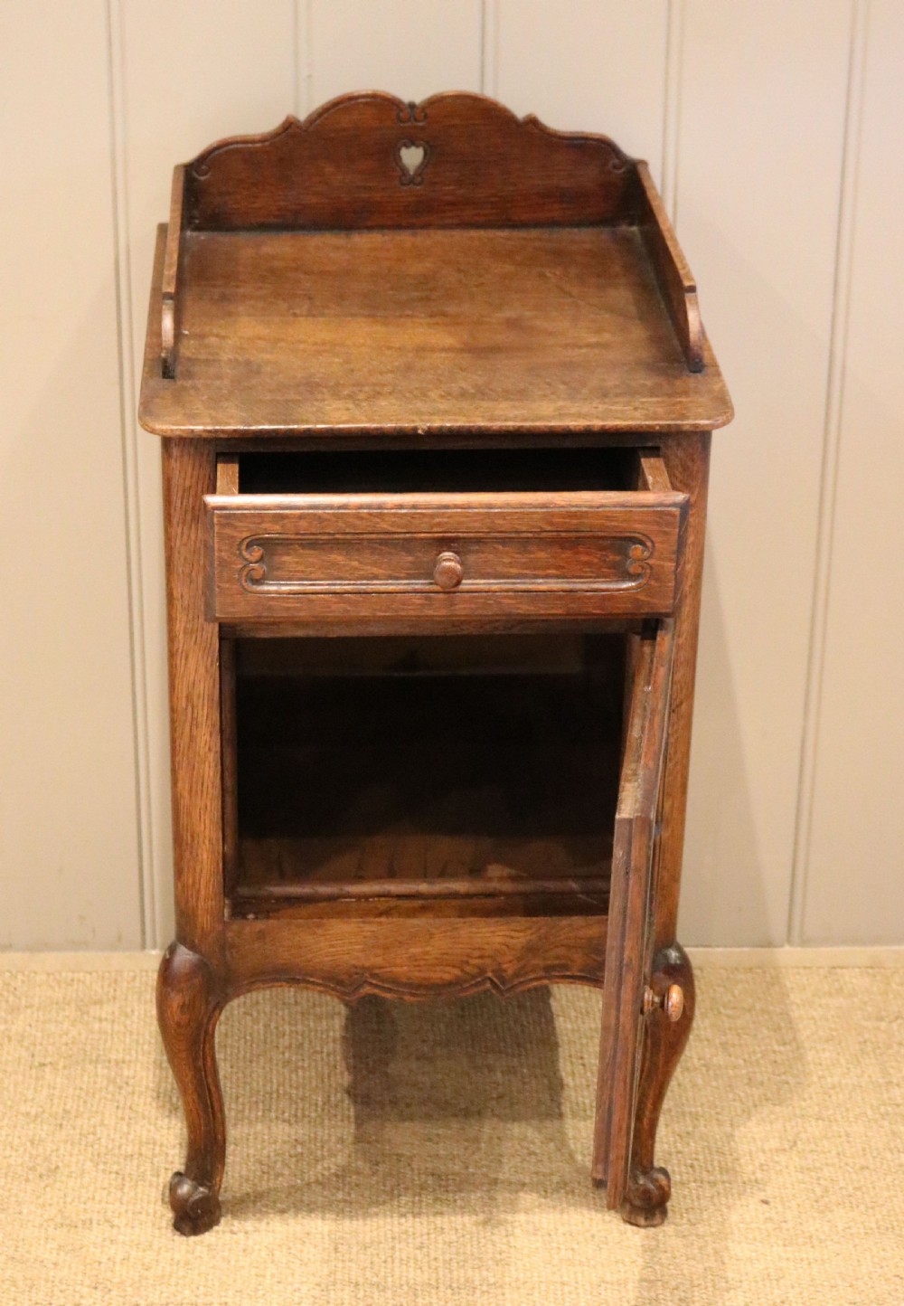 A warm brown solid oak bedside cabinet featuring a heart cut-out gallery, a single drawer, a carved panel door, and cabriole legs. View IV