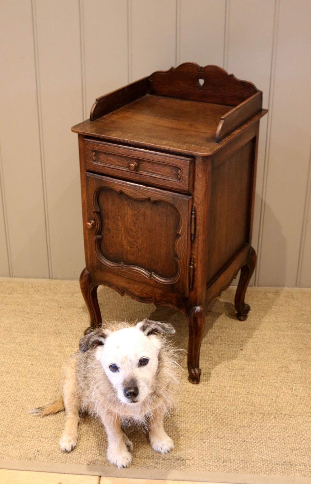 A warm brown solid oak bedside cabinet featuring a heart cut-out gallery, a single drawer, a carved panel door, and cabriole legs. View V