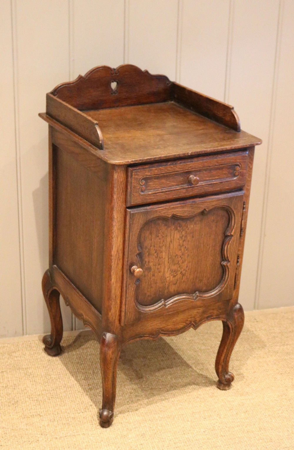 A warm brown solid oak bedside cabinet featuring a heart cut-out gallery, a single drawer, a carved panel door, and cabriole legs. View VI