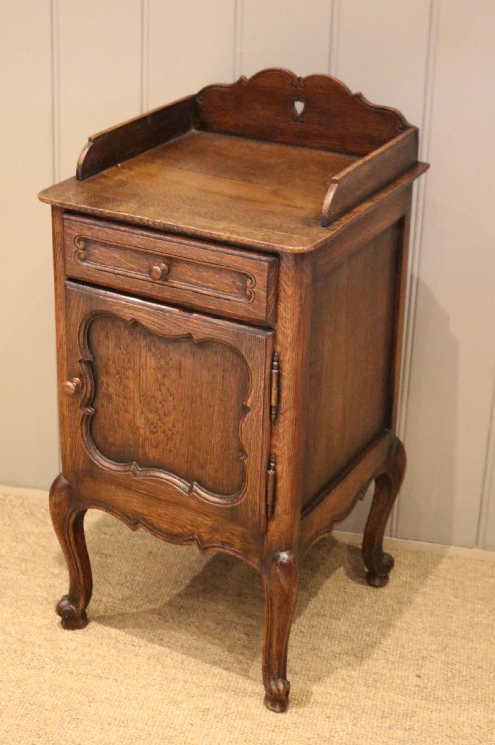 A warm brown solid oak bedside cabinet featuring a heart cut-out gallery, a single drawer, a carved panel door, and cabriole legs. View VII