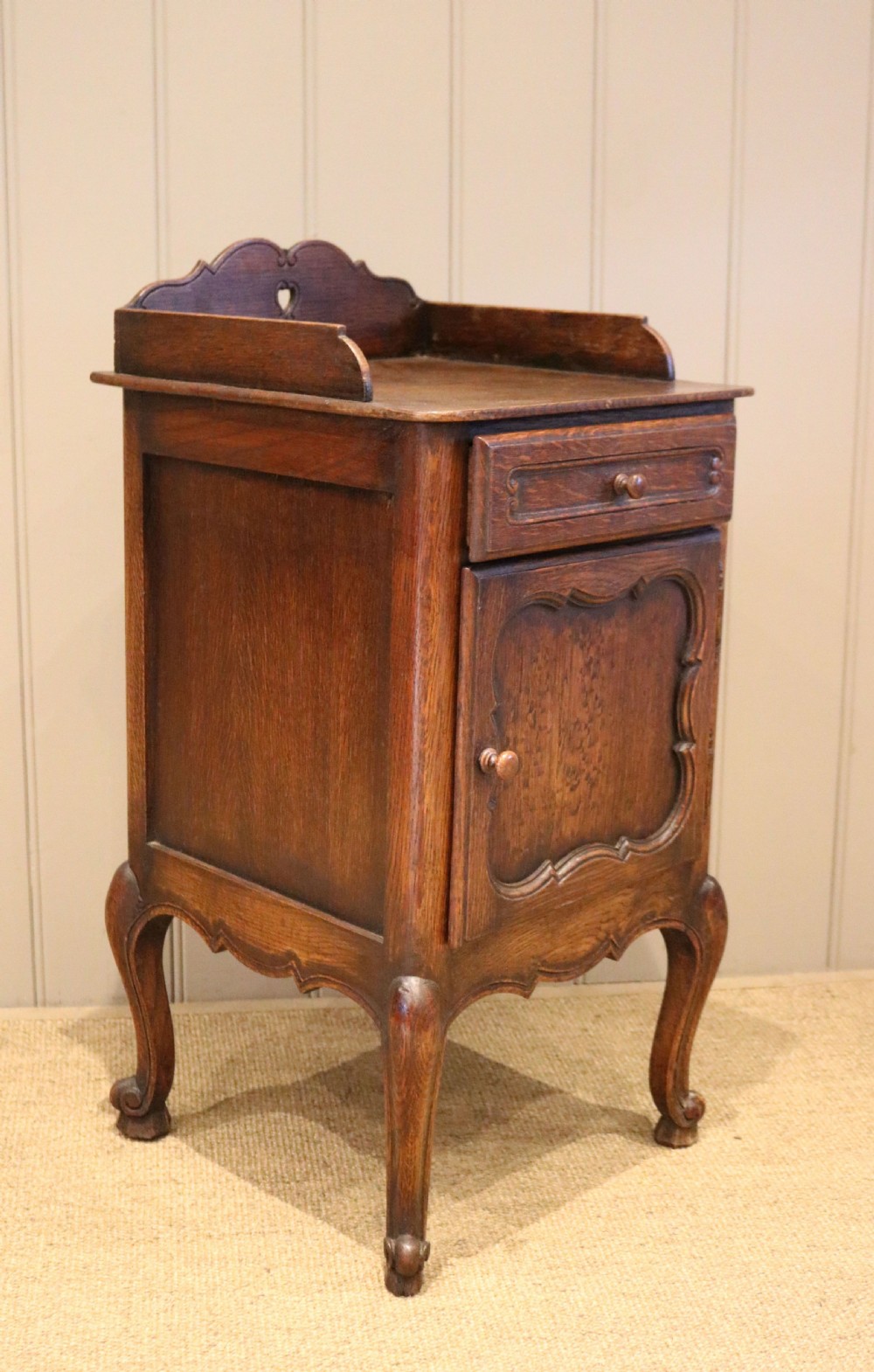 A warm brown solid oak bedside cabinet featuring a heart cut-out gallery, a single drawer, a carved panel door, and cabriole legs. View VIII