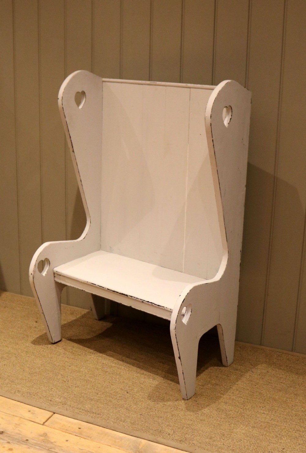 A small off-white painted wooden Arts and Crafts style high-backed settle featuring heart cut-outs on the side panels.