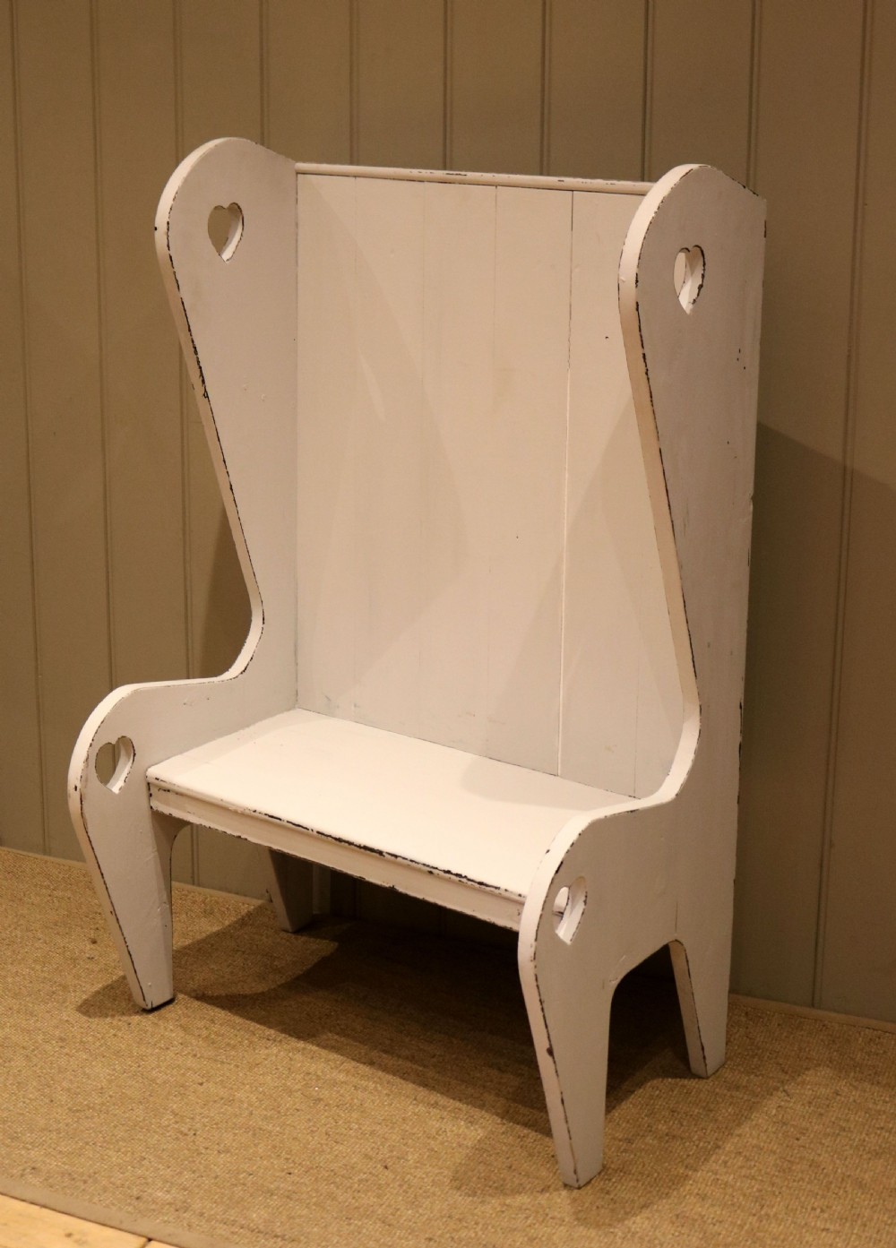 A small off-white painted wooden Arts and Crafts style high-backed settle featuring heart cut-outs on the side panels. View V