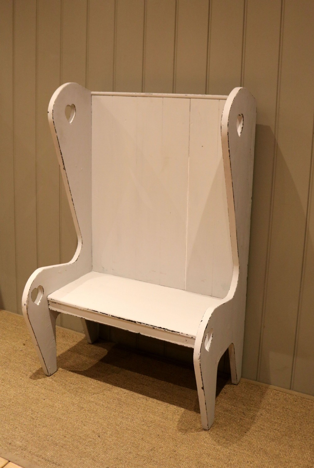 A small off-white painted wooden Arts and Crafts style high-backed settle featuring heart cut-outs on the side panels. View X