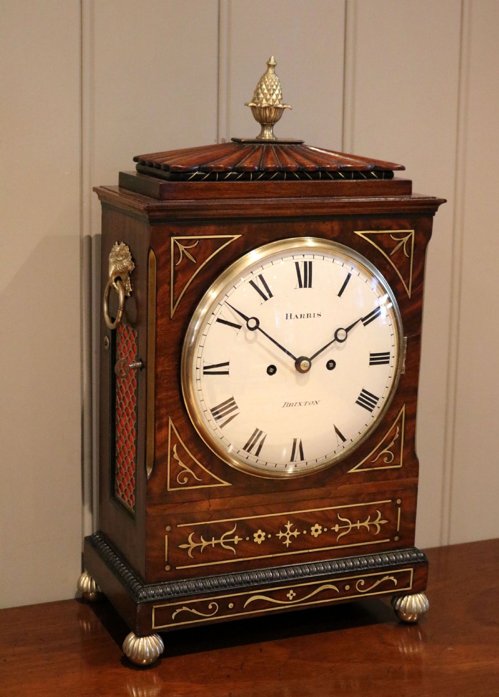 A Regency flame mahogany bracket clock with detailed brass inlay, a white circular dial signed Harris Brixton, side ring handles, and a brass pineapple finial.