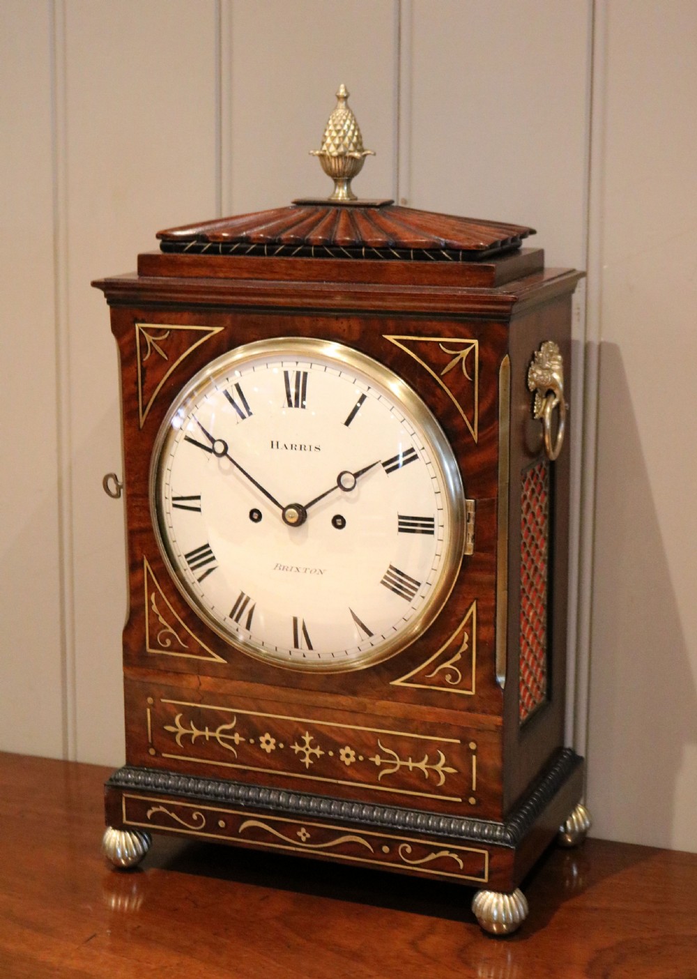 A Regency flame mahogany bracket clock with detailed brass inlay, a white circular dial signed Harris Brixton, side ring handles, and a brass pineapple finial. View II