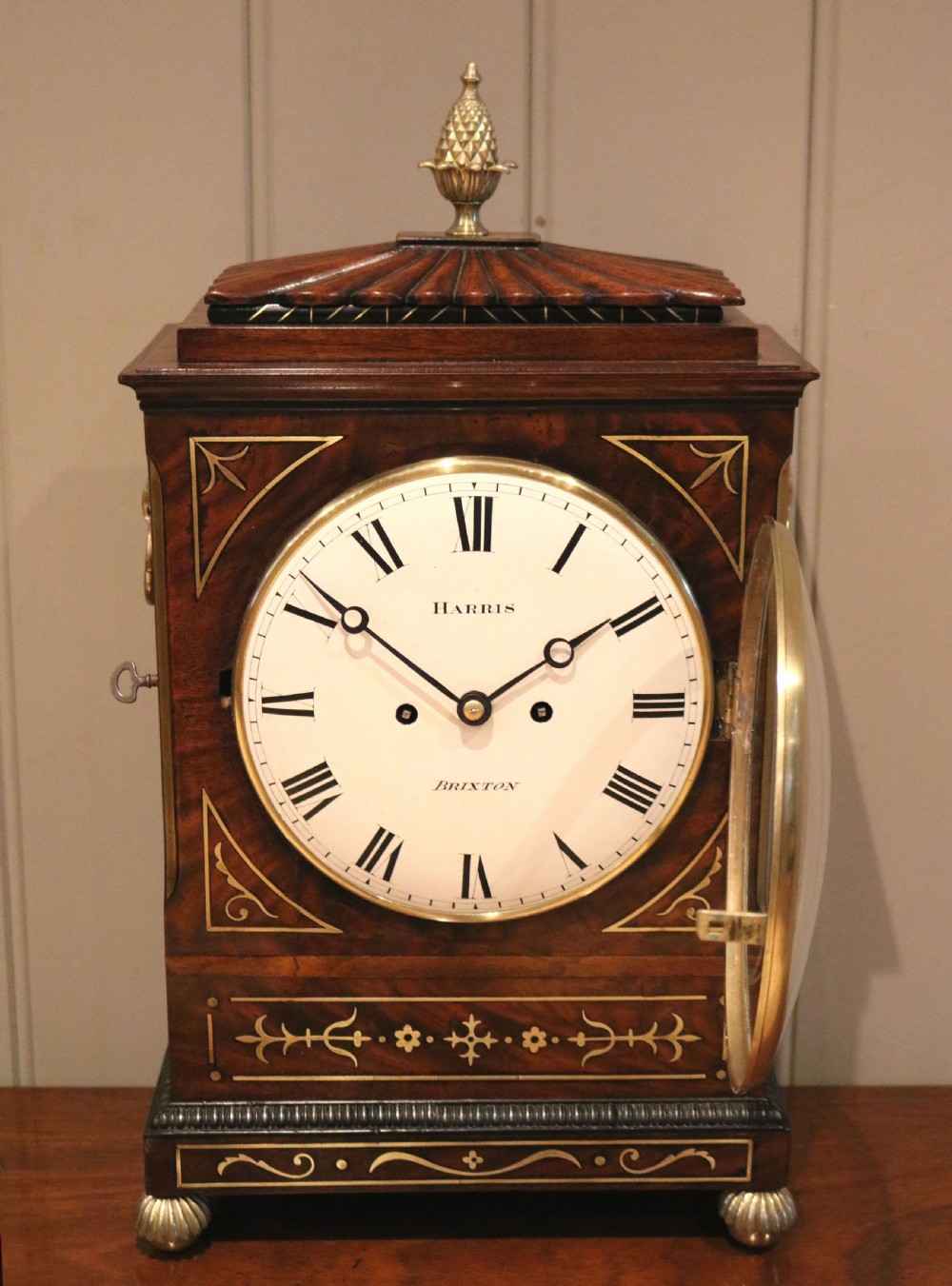 A Regency flame mahogany bracket clock with detailed brass inlay, a white circular dial signed Harris Brixton, side ring handles, and a brass pineapple finial. View III