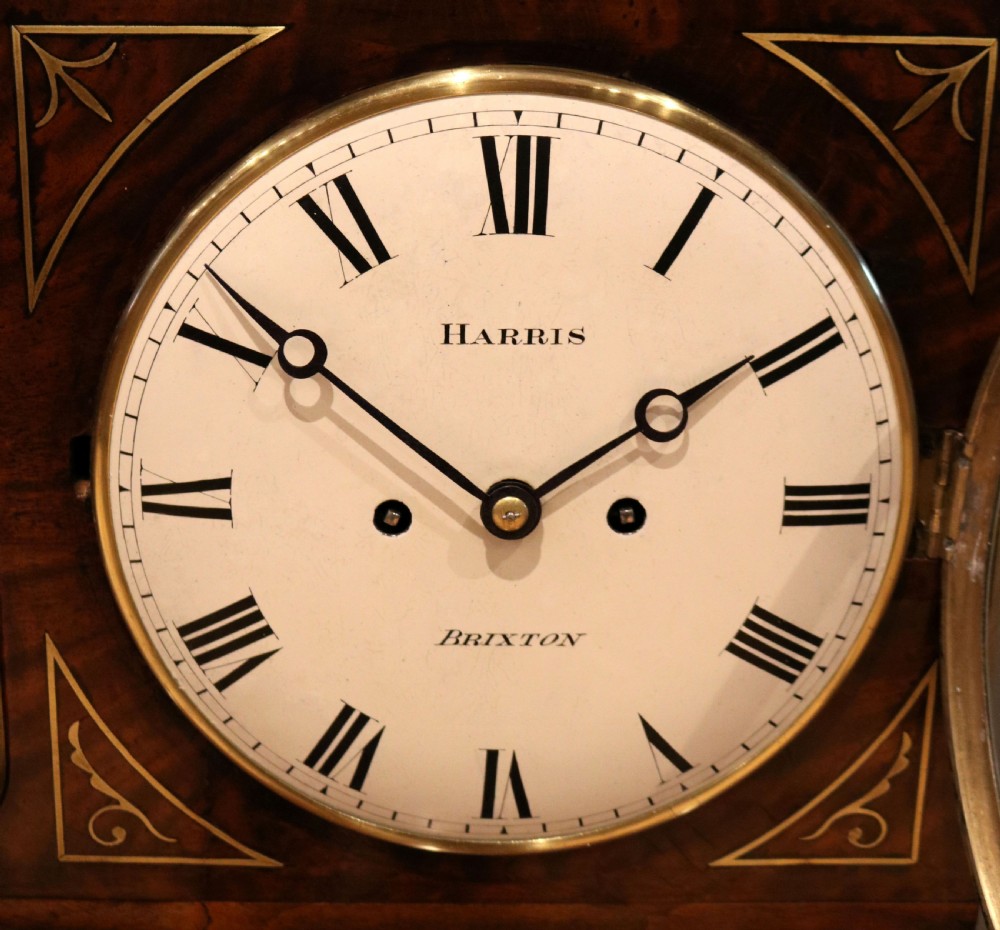 A Regency flame mahogany bracket clock with detailed brass inlay, a white circular dial signed Harris Brixton, side ring handles, and a brass pineapple finial. View IV