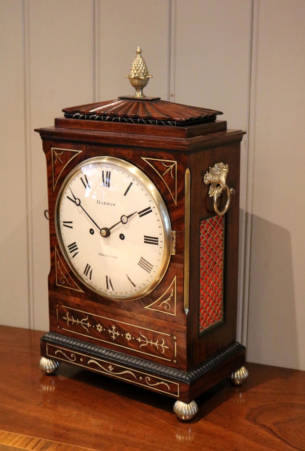 A Regency flame mahogany bracket clock with detailed brass inlay, a white circular dial signed Harris Brixton, side ring handles, and a brass pineapple finial. View IX
