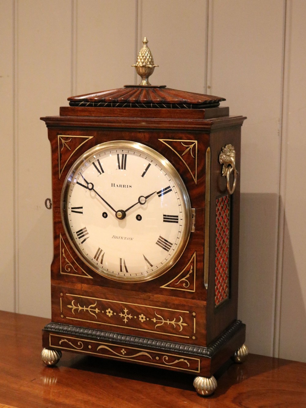 A Regency flame mahogany bracket clock with detailed brass inlay, a white circular dial signed Harris Brixton, side ring handles, and a brass pineapple finial. View XII