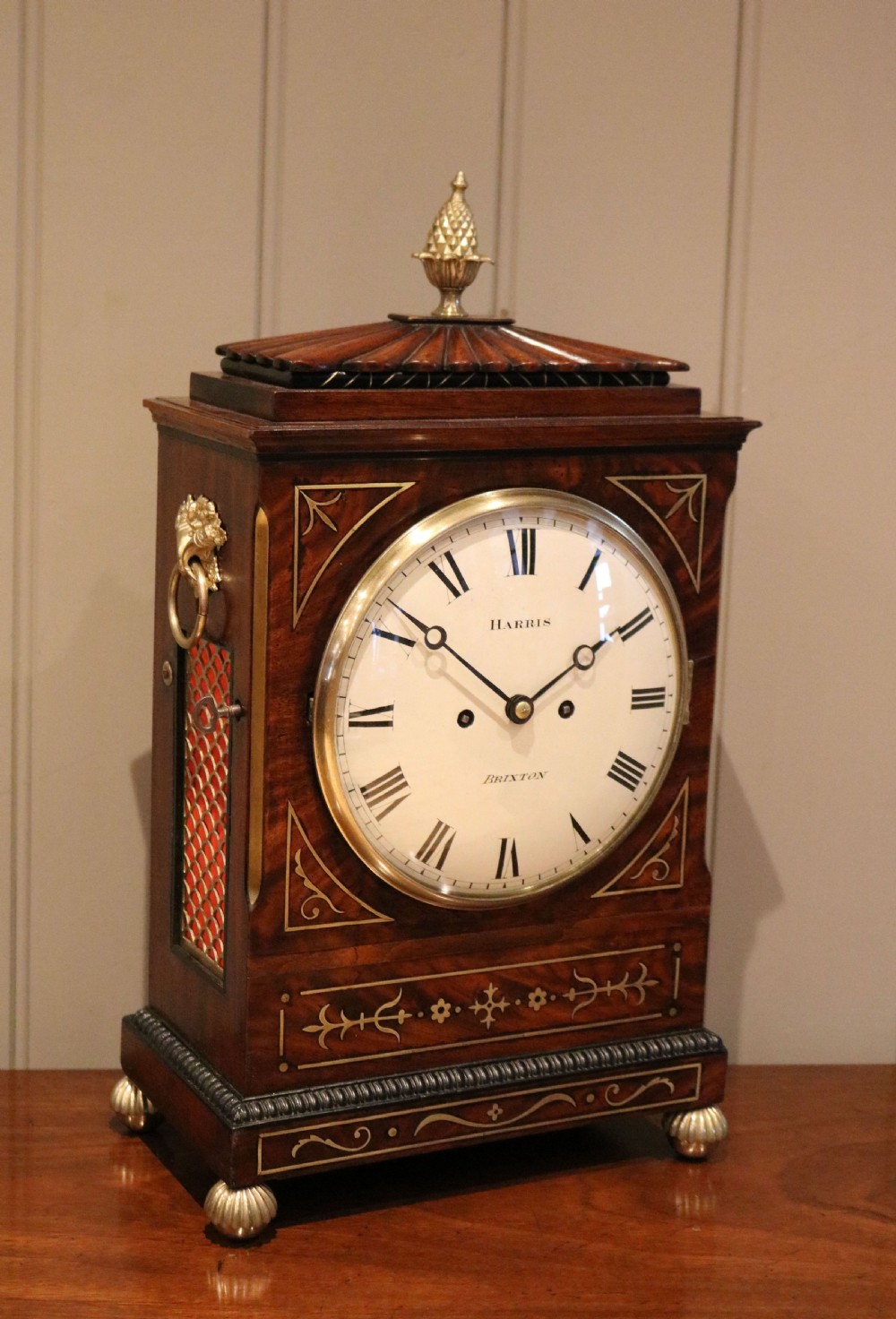 A Regency flame mahogany bracket clock with detailed brass inlay, a white circular dial signed Harris Brixton, side ring handles, and a brass pineapple finial. View XIII