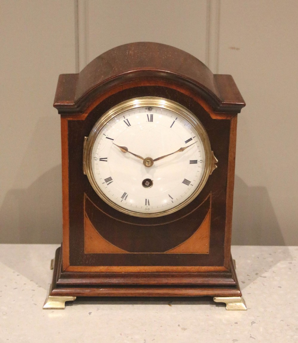 An arched mahogany mantel clock featuring geometric satinwood inlay, a white dial with Roman numerals, a brass bezel, and small brass feet.
