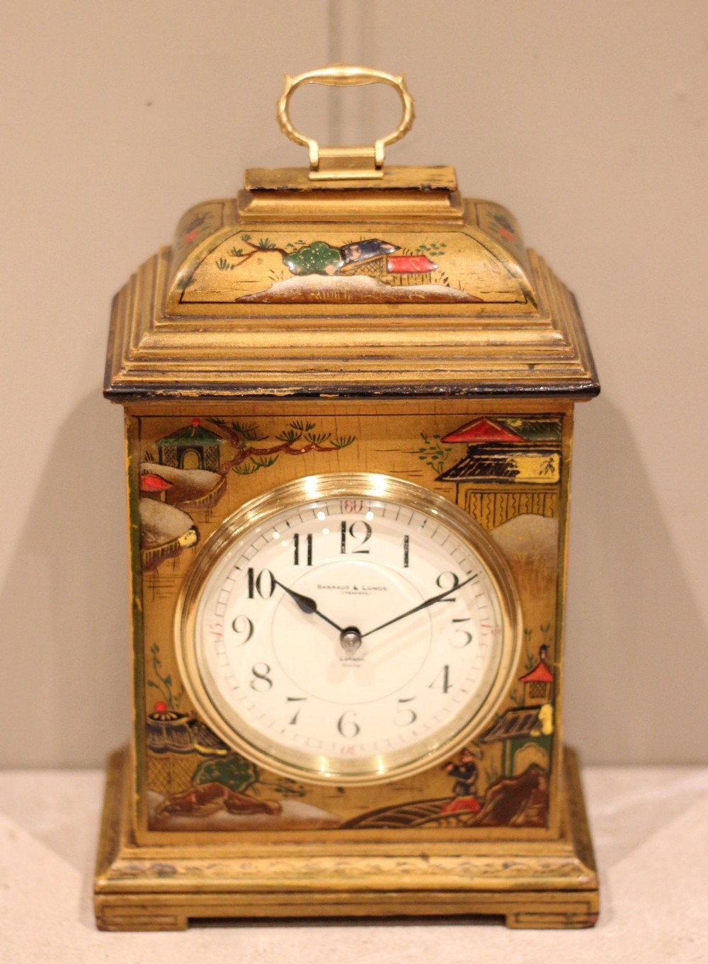A small rectangular Chinoiserie lacquered mantel clock decorated with hand-painted gold and polychrome Asian landscapes, featuring a white dial and top brass handle.