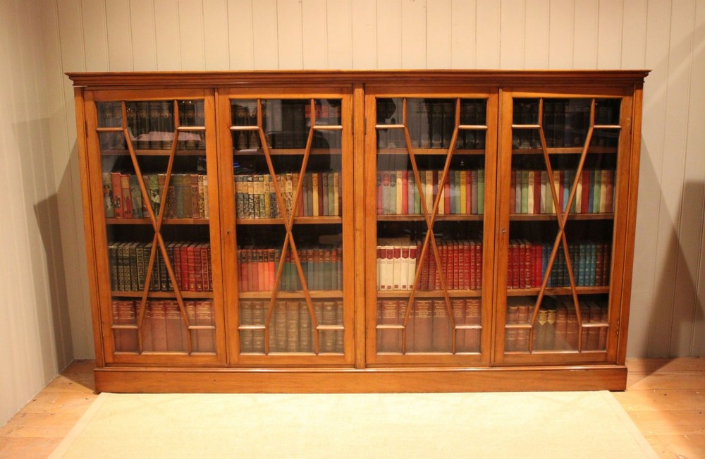 A wide four-door Edwardian walnut library bookcase featuring geometric astragal glazed doors and a plinth base, filled with books.