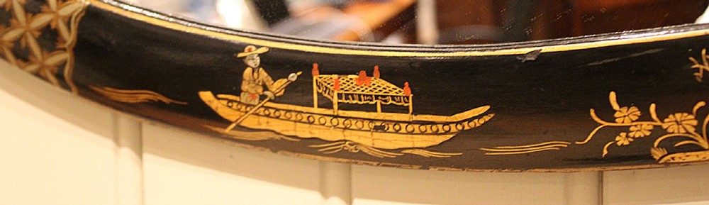 An oval wall mirror featuring a black lacquered wood frame decorated with intricate gold Chinoiserie landscapes, figures, and geometric patterns. View VI