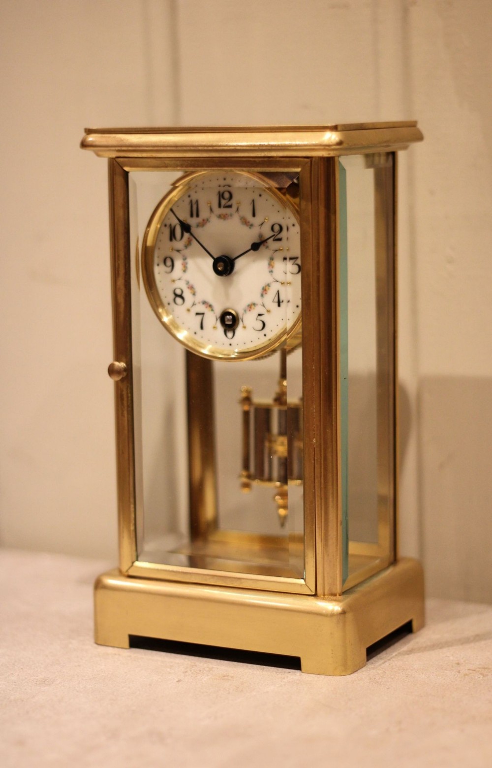 A rectangular brass four glass mantel clock featuring bevelled glass panels, a white enamel dial with floral garland decoration, and a visible hanging pendulum.