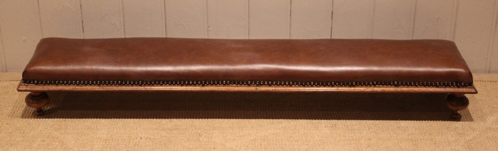 A long, low Victorian footstool upholstered in warm brown leather with brass studding along the lower edge, supported by short turned walnut feet.