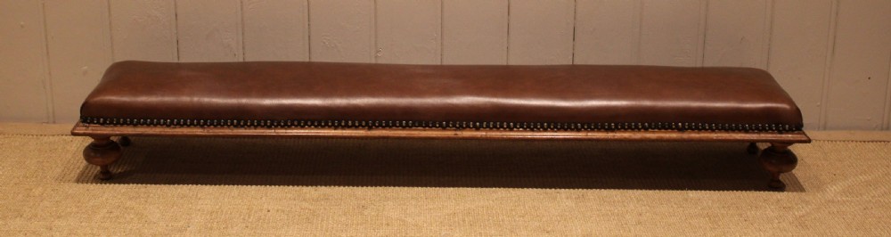 A long, low Victorian footstool upholstered in warm brown leather with brass studding along the lower edge, supported by short turned walnut feet. View III