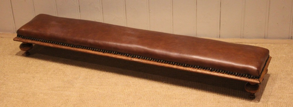 A long, low Victorian footstool upholstered in warm brown leather with brass studding along the lower edge, supported by short turned walnut feet. View IV