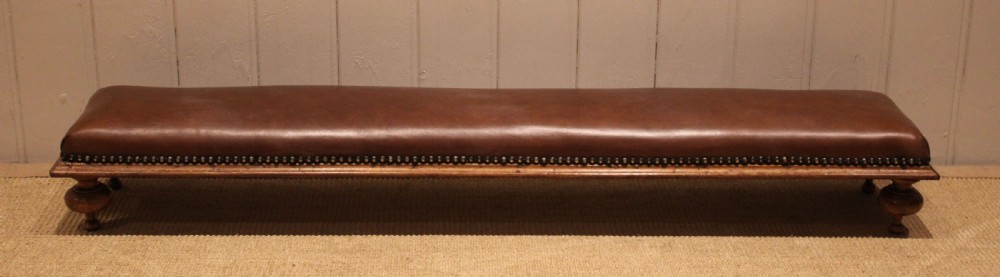 A long, low Victorian footstool upholstered in warm brown leather with brass studding along the lower edge, supported by short turned walnut feet. View V